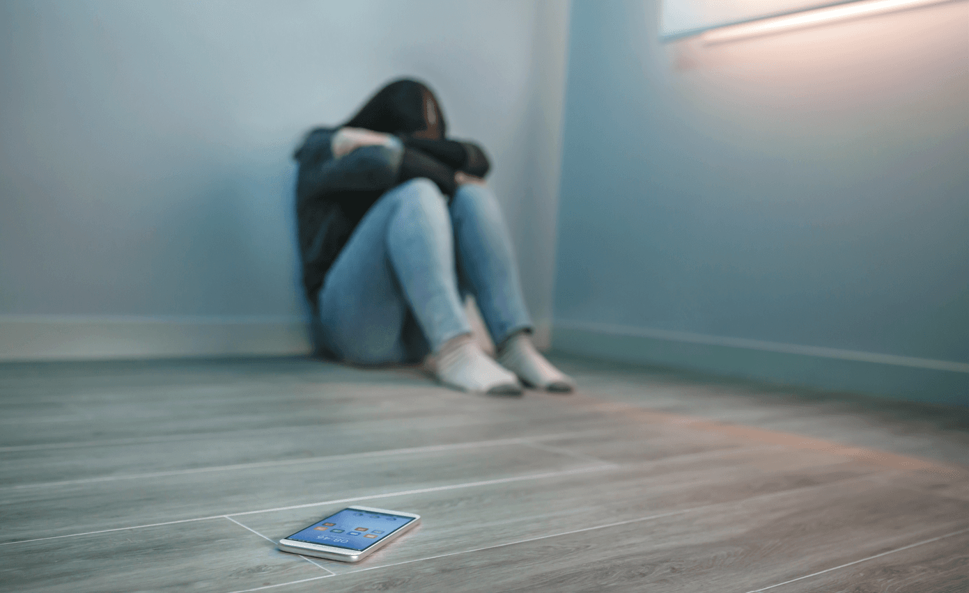 Dziewczynka skulona pod ścianą na podłodze, niedaleko niej leży telefon. 