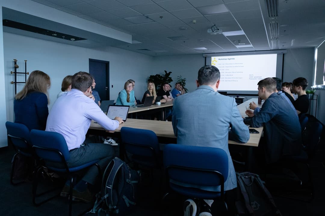 Zdjęcie przedstawia grupę uczestników warsztatu siedzących przy dużym stole konferencyjnym. Na ekranie projektora widoczna jest agenda spotkania zatytułowanego „Workshop Agenda”. Uczestnicy są skupieni na dyskusji, niektórzy korzystają z laptopów.