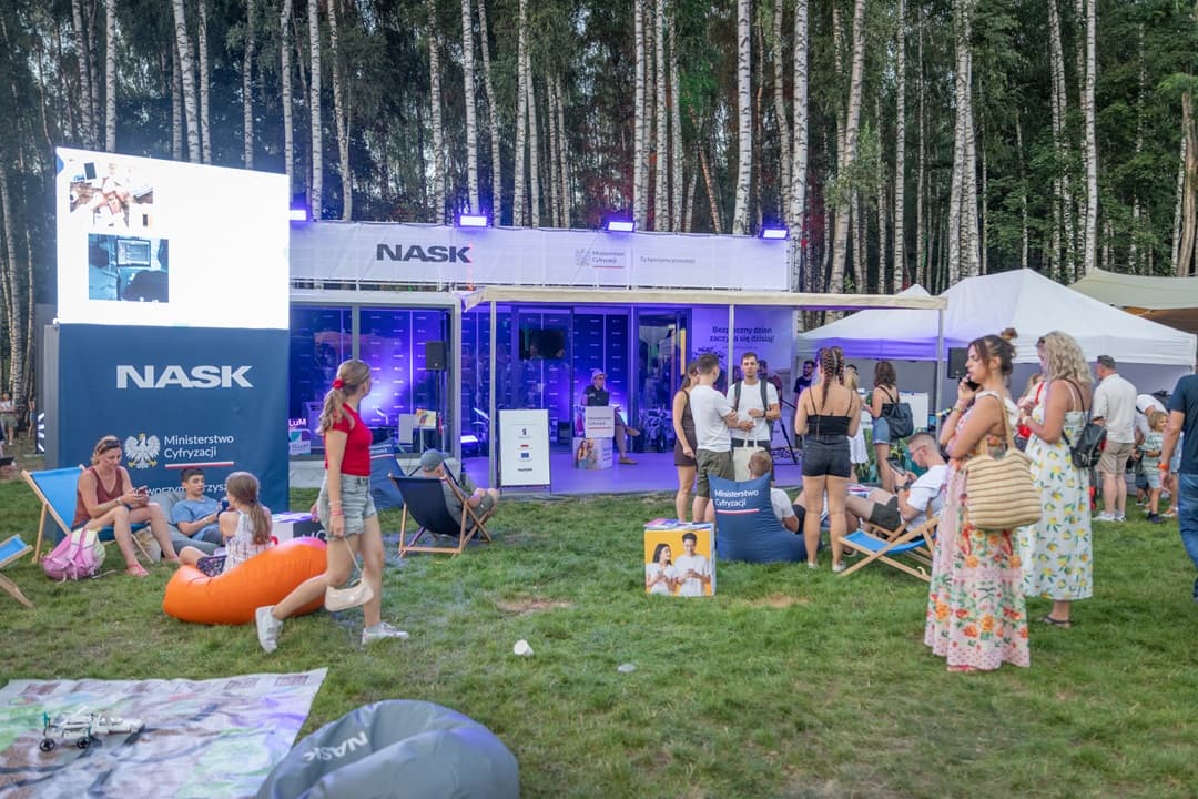 Plenerowe wydarzenie w lesie z widocznym stoiskiem NASK pośrodku. Po lewej stronie duży ekran wyświetlający obrazy. Przed stoiskiem ludzie siedzą na pufach i leżakach, rozmawiają i odpoczywają. W tle wysokie drzewa.