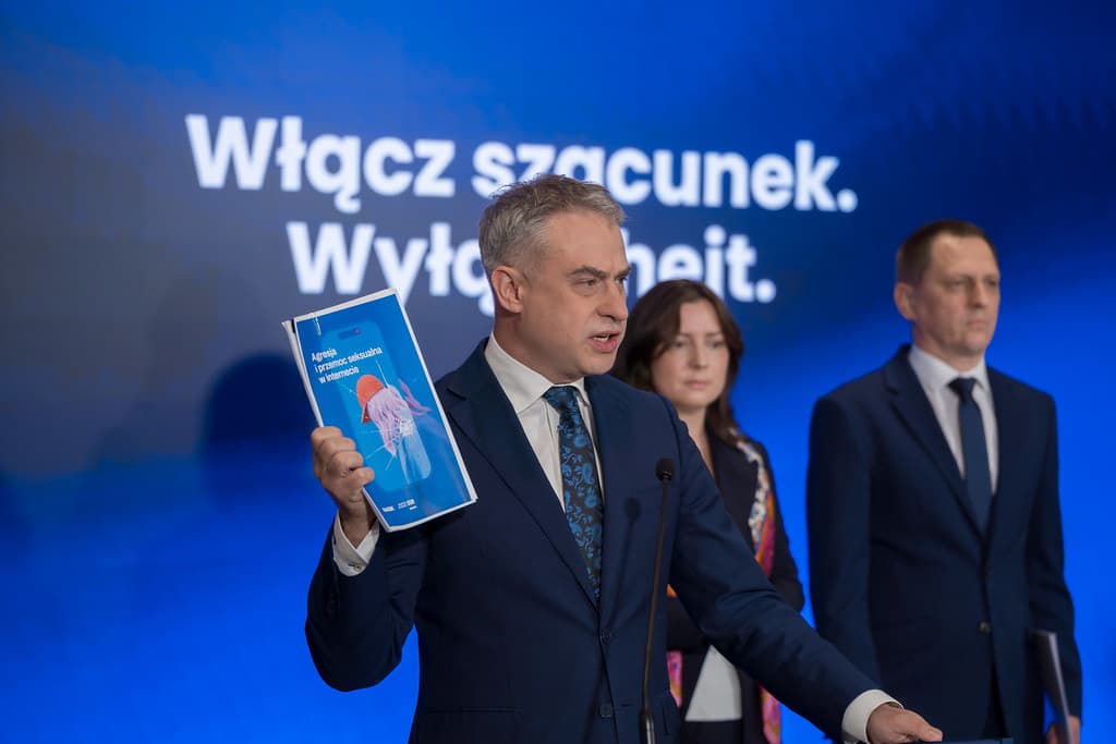 Wicepremier i minister cyfryzacji Krzysztof Gawkowski prezentuje raport NASK „Agresja i przemoc seksualna w internecie” podczas konferencji inaugurującej kampanię „Włącz szacunek. Wyłącz hejt”. W tle widać niebieski ekran z hasłem kampanii. Obok ministra stoją przedstawiciele NASK i Ministerstwa Cyfryzacji.