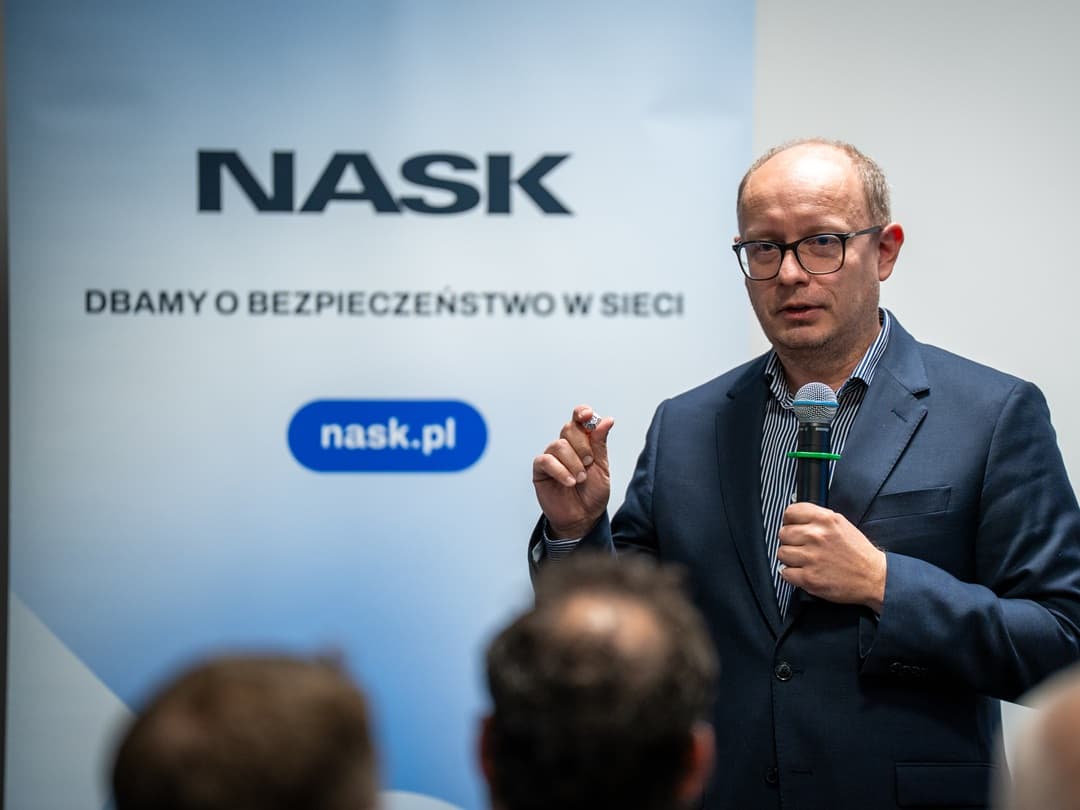 Radosław Nielek przemawia do publiczności, trzymając mikrofon. W tle znajduje się baner z logo NASK oraz napisem „Dbamy o bezpieczeństwo w sieci” i adresem strony „nask.pl”.