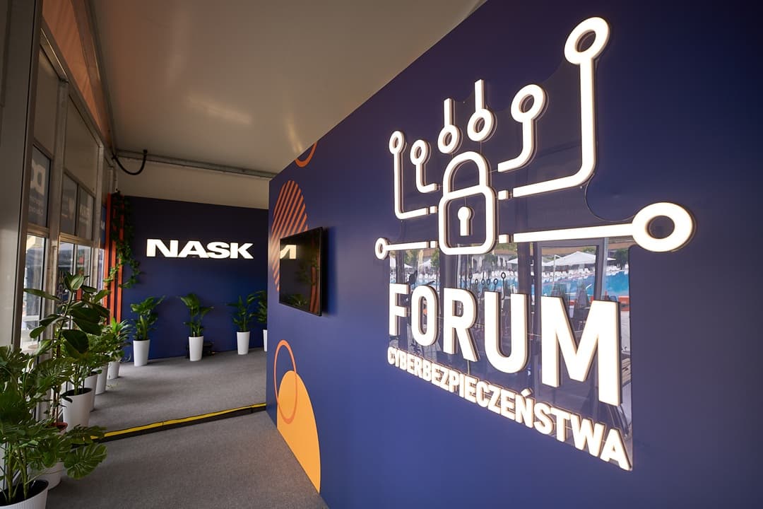 Wnętrze stoiska z ciemnoniebieskimi ścianami ozdobionymi logotypami. Po prawej stronie duży podświetlany napis „Forum Cyberbezpieczeństwa” z ikoną kłódki i symbolami sieci. W głębi widoczny podświetlany napis „NASK” oraz rośliny w białych donicach ustawione wzdłuż ściany.