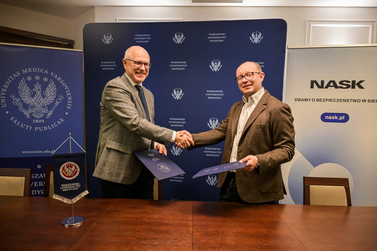 Na zdjęciu prof. Rafał Krenke, rektor Warszawskiego Uniwersytetu Medycznego, oraz Radosław Nielek, dyrektor NASK, ściskają sobie dłonie podczas oficjalnego podpisania umowy o współpracy. W tle widoczne są banery z logo Warszawskiego Uniwersytetu Medycznego oraz NASK. Obaj mężczyźni uśmiechają się i trzymają teczki z dokumentami.