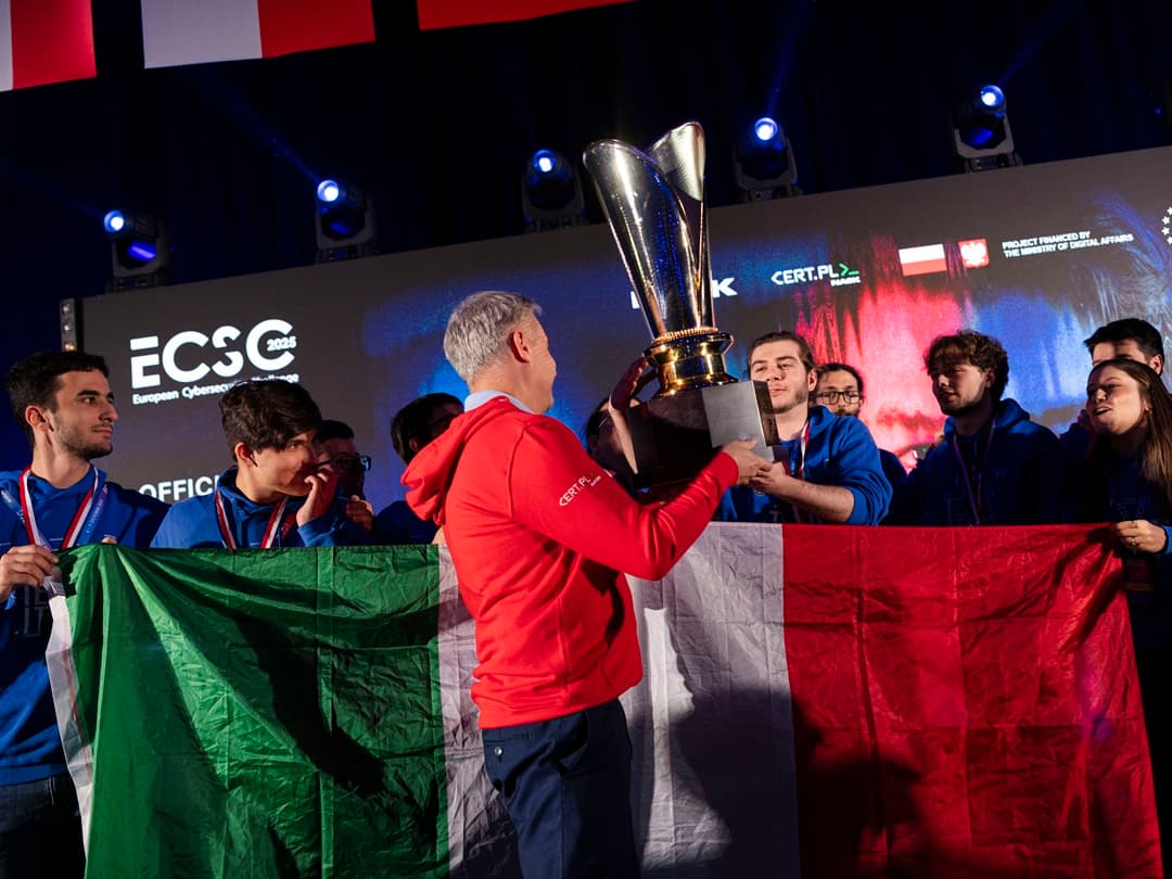 Minister Cyfryzacji Krzysztof Gawkowski wręcza puchar zwycięskiej drużynie z Włoch podczas finału European Cybersecurity Challenge 2025. Członkowie zespołu ubrani w niebieskie bluzy trzymają dużą włoską flagę i cieszą się ze zwycięstwa.
