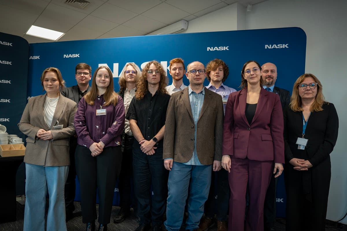 Grupa uczestników i organizatorów zawodów OSINT CTF NASK pozuje do wspólnego zdjęcia na tle niebieskiej ścianki z logo NASK.