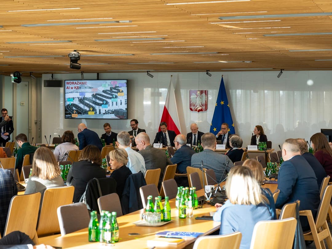 Zdjęcie przedstawia salę konferencyjną z wieloma osobami siedzącymi przy stołach i słuchającymi panelu ekspertów umieszczonych przy długim stole na przodzie. Na ścianie za panelistami widoczne są flagi Polski i Unii Europejskiej oraz herb Polski. Na ekranie wyświetlona jest prezentacja dotycząca wdrażania sztucznej inteligencji. Na stołach stoją liczne zielone butelki z wodą.