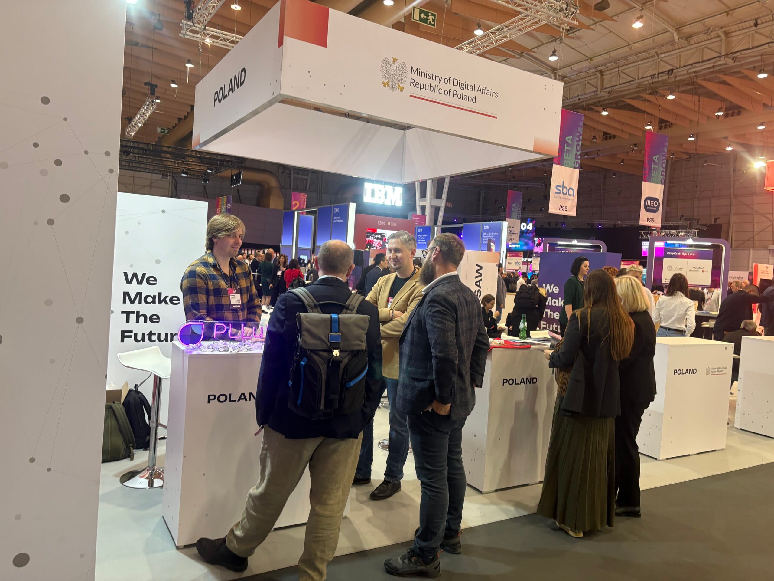 Stoisko Polski podczas międzynarodowych targów technologicznych, oznaczone logotypem Ministerstwa Cyfryzacji i hasłem „We Make The Future”. Uczestnicy rozmawiają przy ladach wystawowych.