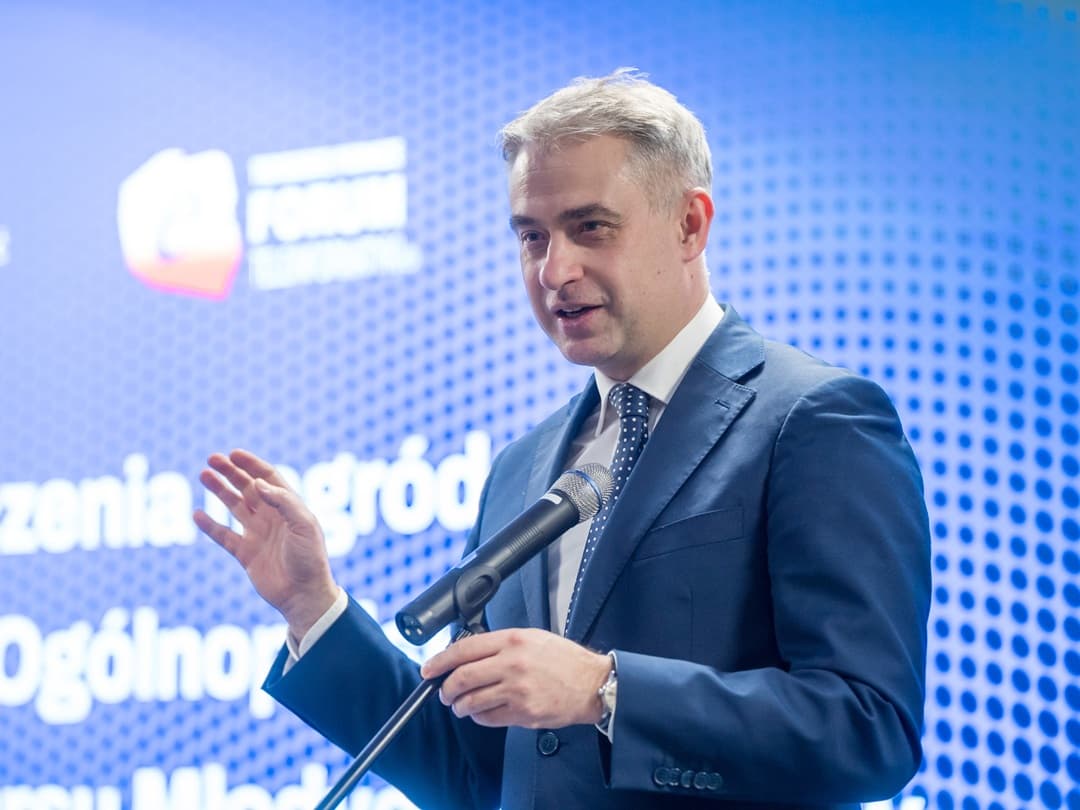 Krzysztof Gawkowski w granatowym garniturze przemawiający do mikrofonu na stojaku podczas konferencji. Jedną rękę ma uniesioną w geście mówienia. W tle niebieski ekran z rozmytym napisem i graficznym wzorem kropek.