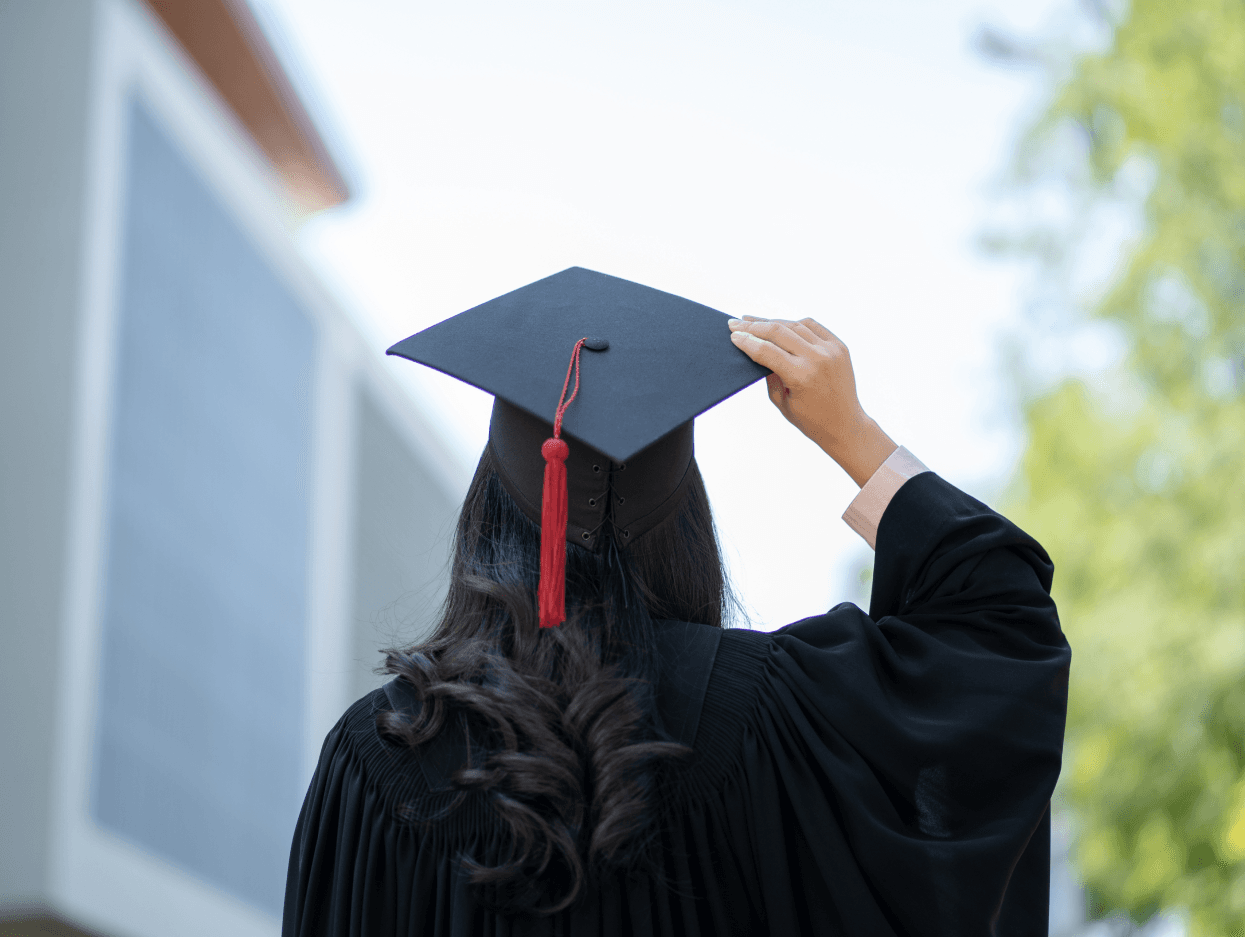 Stojąca tyłem dziewczyna w todze przytrzymuje jedną ręką biret studencki