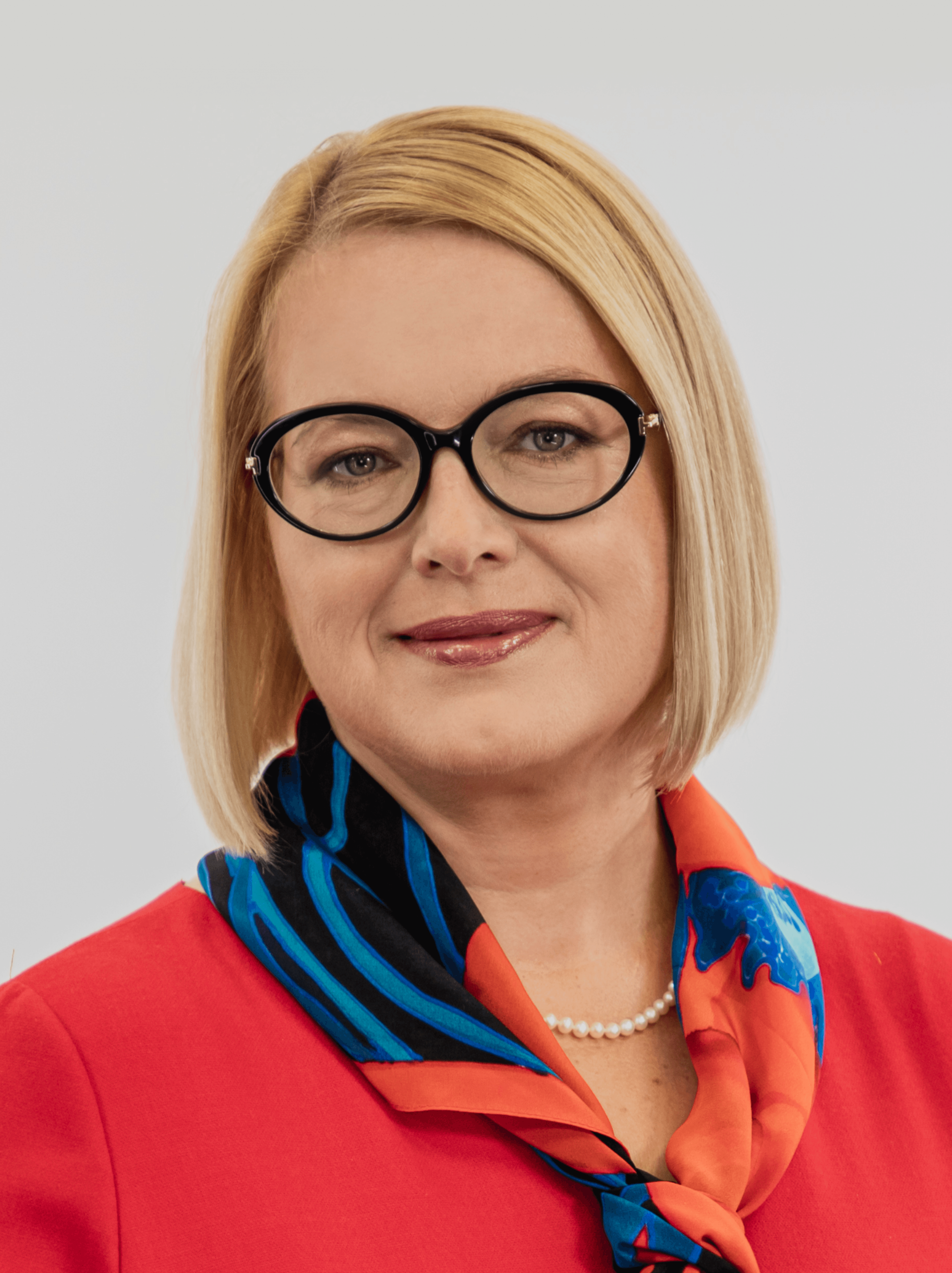 Półportret. Lekko uśmiechnięta blondynka w okularach i czerwonej marynarce patrzy w obiektyw. Magdalena Tarczewska-Szymanska
