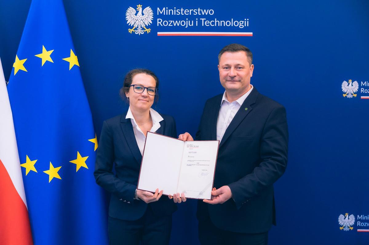 Dr Marta Wachowicz i minister Krzysztof Paszyk podczas wręczenia aktu powołania na stanowisko prezesa Polskiej Agencji Kosmicznej. W tle logotyp Ministerstwa Rozwoju i Technologii oraz flagi Polski i Unii Europejskiej.