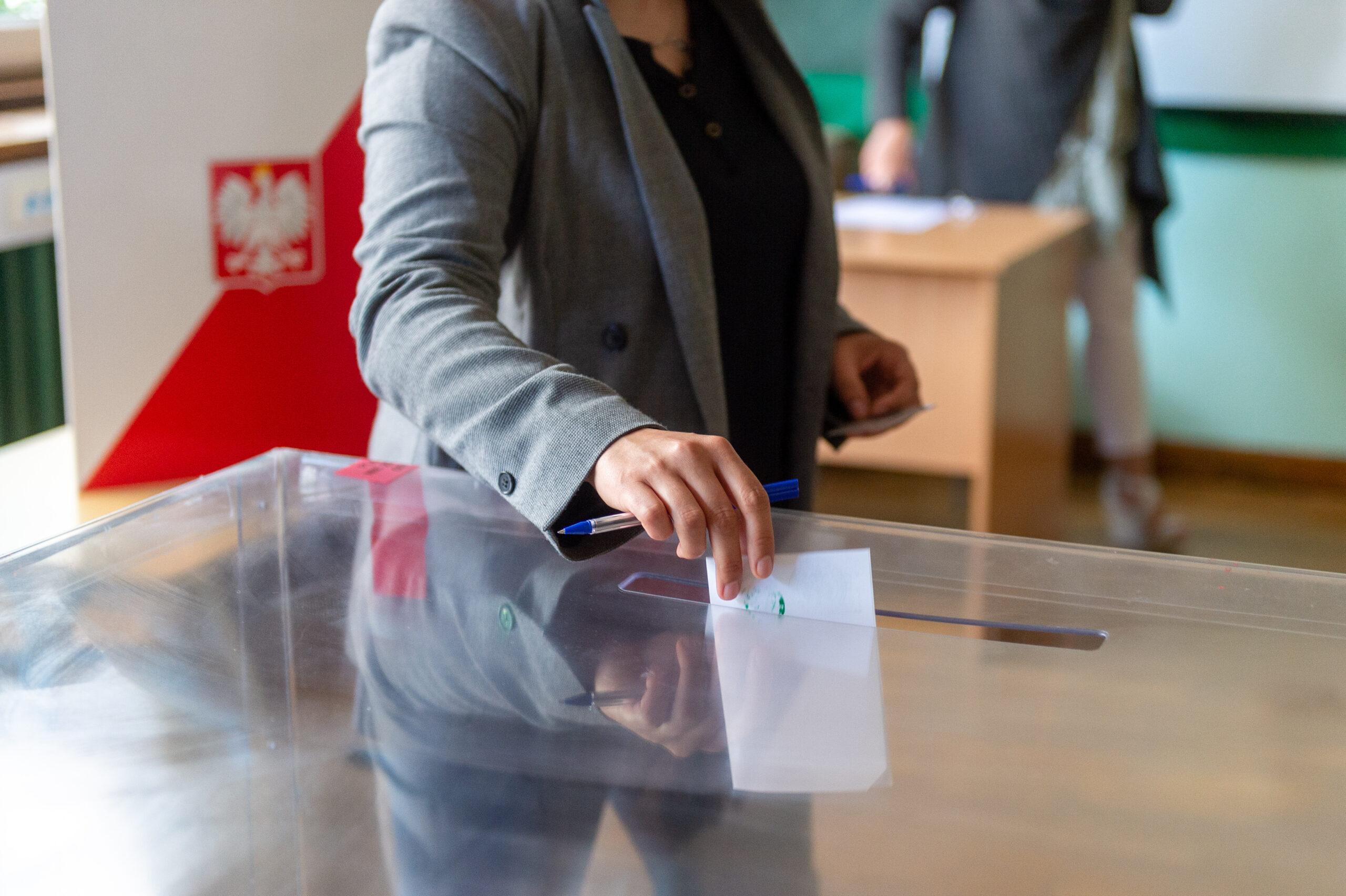 Kobieta, pokazana na zdjęciu od ramion w dół, stojąca przy urnie w lokalu wyborczym, wrzucająca kopertę.