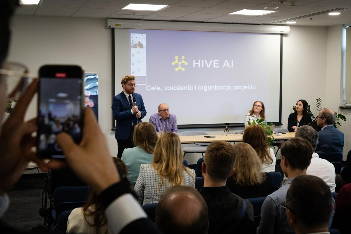 Wiceminister cyfryzacji Dariusz Standerski przemawia podczas konferencji dotyczącej projektu HIVE AI. W tle na ekranie widoczne są slajdy prezentacji z tytułem „Cele, założenia i organizacja projektu”. Przy stole prezydialnym siedzą Radosław Nielek, Agnieszka Karlińska. W sali znajduje się licznie zgromadzona publiczność, a jedno z przemówień jest rejestrowane telefonem przez uczestnika.