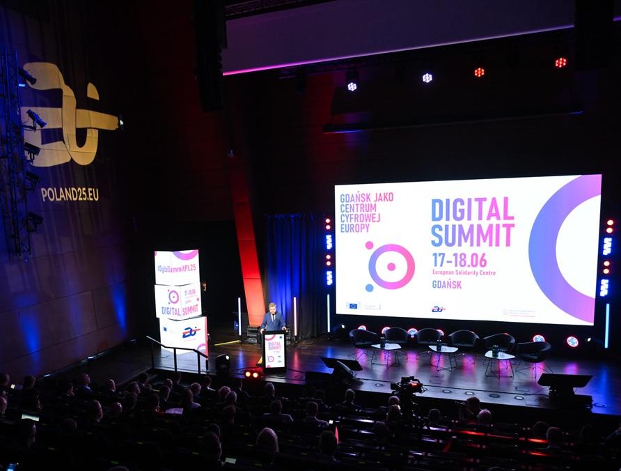 Wiceminister cyfryzacji Dariusz Standerski przemawia na scenie podczas Digital Summit w Europejskim Centrum Solidarności w Gdańsku. Za nim znajduje się duży ekran z grafiką wydarzenia zawierającą nazwę „DIGITAL SUMMIT”, daty 17–18.06 oraz lokalizację. Na ekranie widoczne są również logotypy i hasło „Gdańsk jako centrum cyfrowej Europy”. Na sali obecna jest publiczność, scena oświetlona jest światłem w odcieniach niebieskiego i fioletowego.