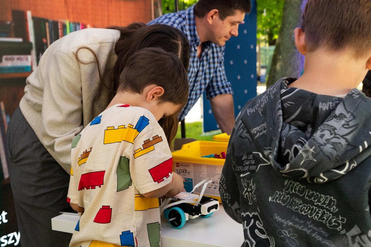 Dzieci uczestniczą w warsztatach z robotyki. Jedno z dzieci, ubrane w koszulkę z kolorowymi klockami, pracuje nad małym robotem z zestawu LEGO Education SPIKE. W tle widać opiekunów wspierających uczestników oraz pojemniki z elementami konstrukcyjnymi.