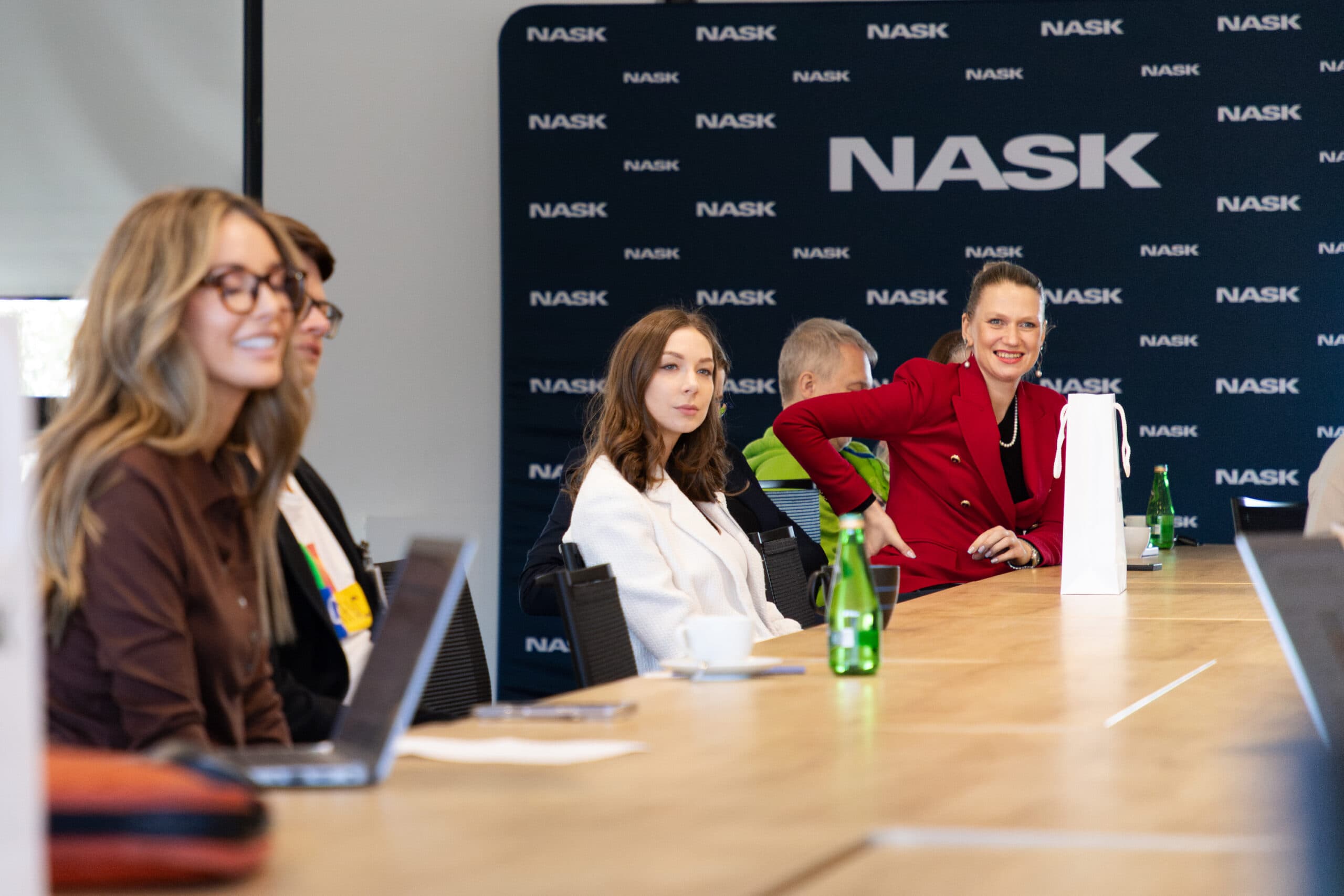 Grupa osób uczestniczy w spotkaniu w siedzibie NASK. Uczestnicy siedzą przy długim stole konferencyjnym, na którym znajdują się laptopy, notatki i butelki wody. W tle widoczne jest logo NASK na granatowej ściance reklamowej. W centrum kadru znajduje się kobieta w czerwonej marynarce, uśmiechająca się w stronę aparatu, obok niej siedzi kobieta w białej marynarce z neutralnym wyrazem twarzy. Po lewej stronie kadru znajduje się kobieta w okularach z uśmiechem na twarzy.