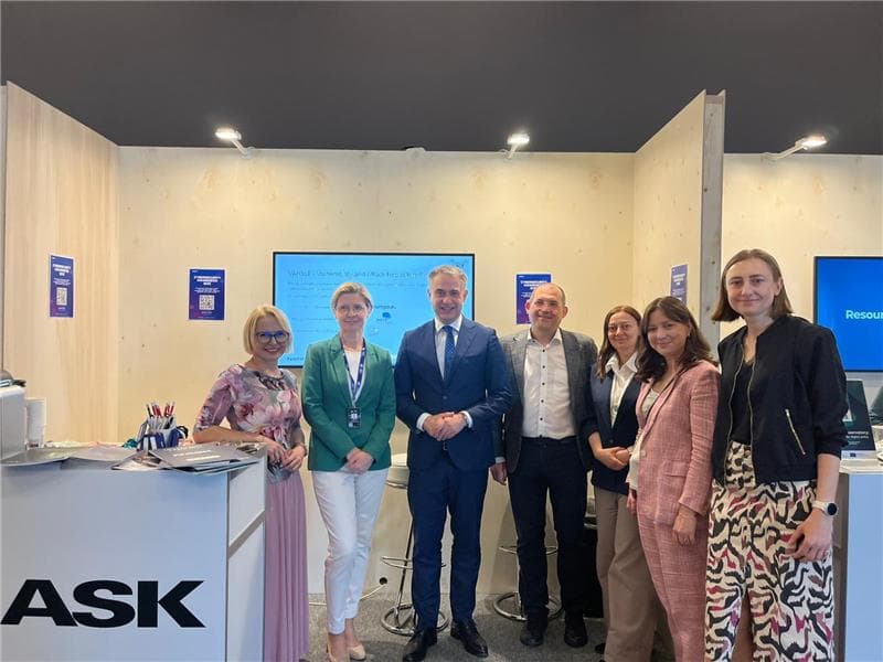 Grupowe zdjęcie siedmiorga osób stojących na stoisku NASK podczas wydarzenia. W tle widoczne są drewniane ścianki, ekran z prezentacją projektu VARIoT oraz plakaty informacyjne. Osoby są uśmiechnięte, ubrane formalnie lub półformalnie, stoją przy ladzie oznaczonej logotypem NASK.