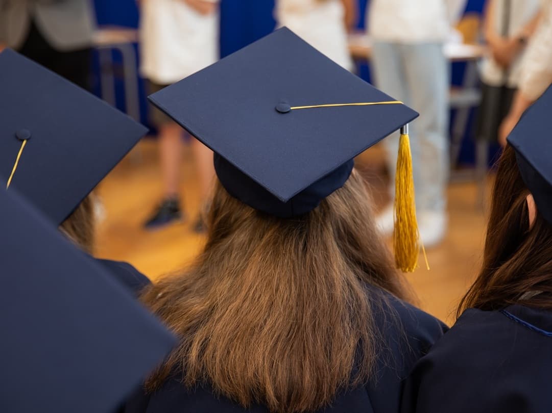 Uczniowie w granatowych biretach z żółtymi chwostami siedzą tyłem podczas szkolnej uroczystości zakończenia roku.