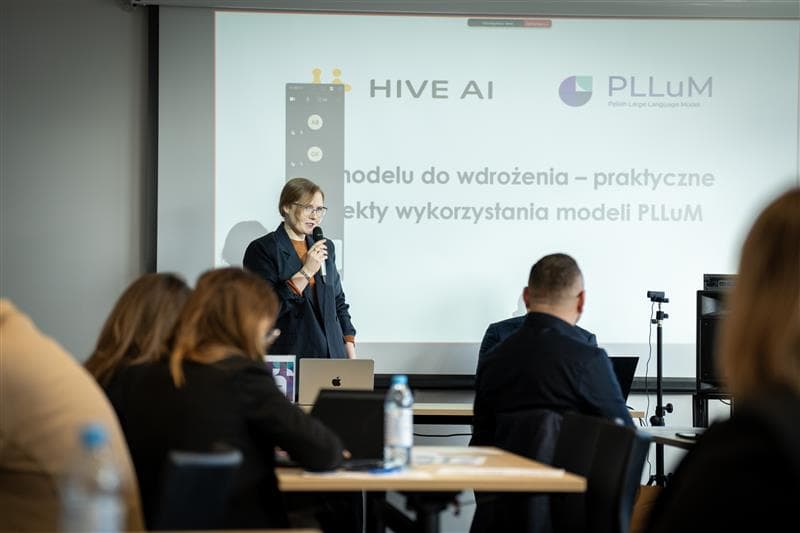 Na zdjęciu Agnieszka Karlińska przemawia do mikrofonu podczas prezentacji w sali konferencyjnej. Za nią widoczny jest ekran z logotypami HIVE AI i PLLuM oraz slajd prezentacji dotyczący wykorzystania modeli językowych. Wokół siedzą uczestnicy spotkania, niektórzy pracują na laptopach.