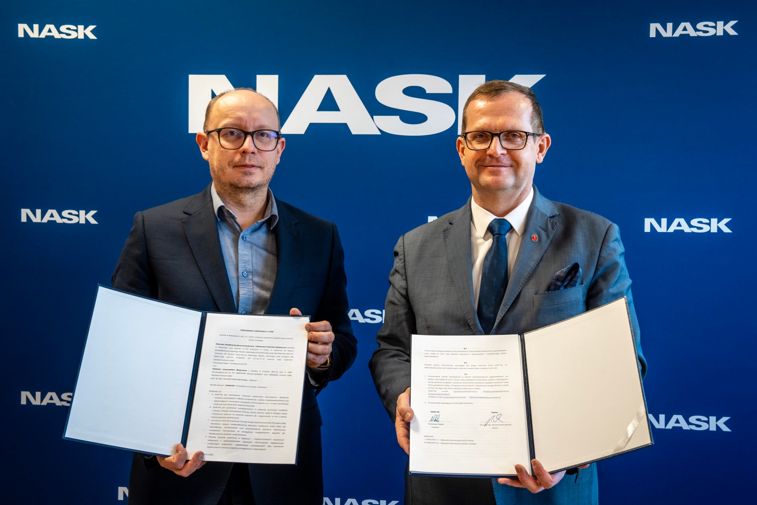 Dwaj mężczyźni stoją przed ścianą z logotypami NASK, trzymając otwarte teczki z podpisanym porozumieniem o współpracy.