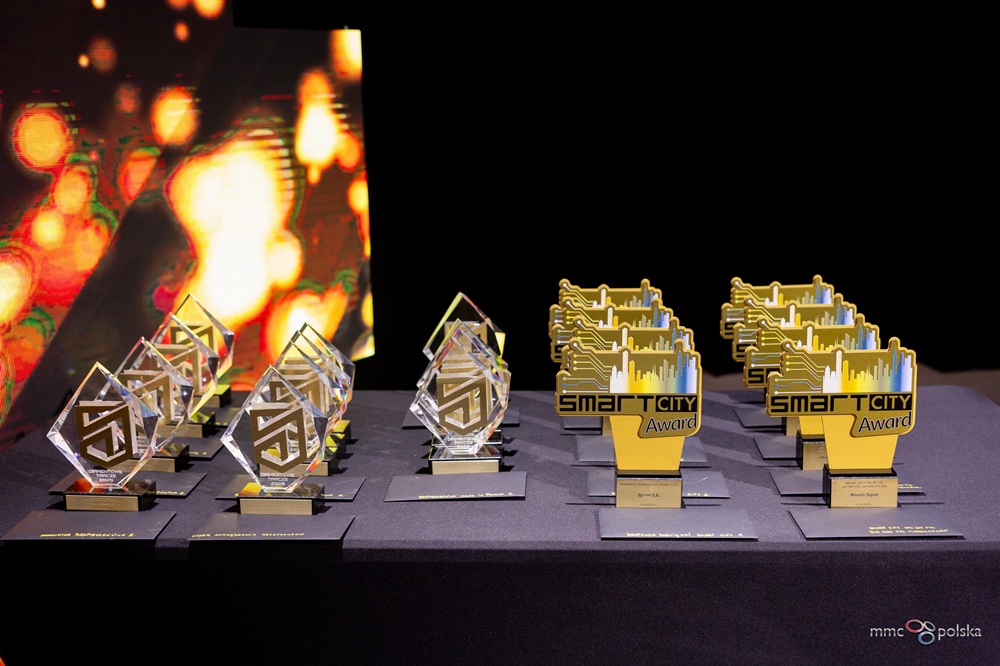 Stół pokryty czarnym obrusem, na którym ustawiono wiele nagród. Po lewej stronie znajdują się eleganckie statuetki w kształcie rombu wykonane z przezroczystego szkła. Po prawej stronie stoją żółto-niebieskie nagrody z motywem miasta i obwodów elektronicznych. W tle widać kolorowe, rozmyte światła.