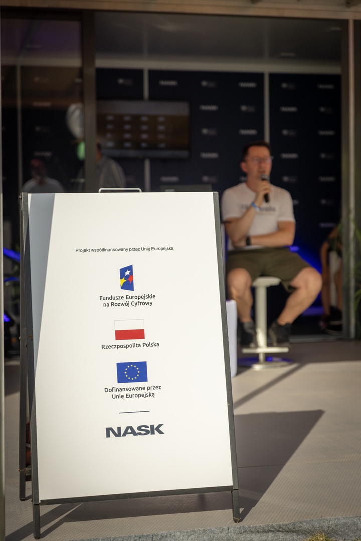 Tablica informacyjna na sztaludze z tekstem o współfinansowaniu projektu przez Unię Europejską oraz logo NASK. W tle osoba siedząca na krześle, trzymająca mikrofon.