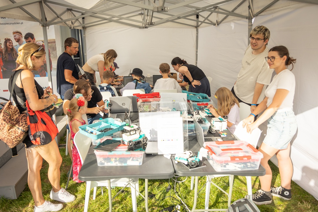 Grupa dzieci i dorosłych uczestniczy w warsztatach edukacyjnych na świeżym powietrzu pod namiotem. Na stołach znajdują się zestawy do budowy robotów i urządzeń technicznych. Dzieci składają i programują urządzenia, a dorośli im pomagają i nadzorują pracę.