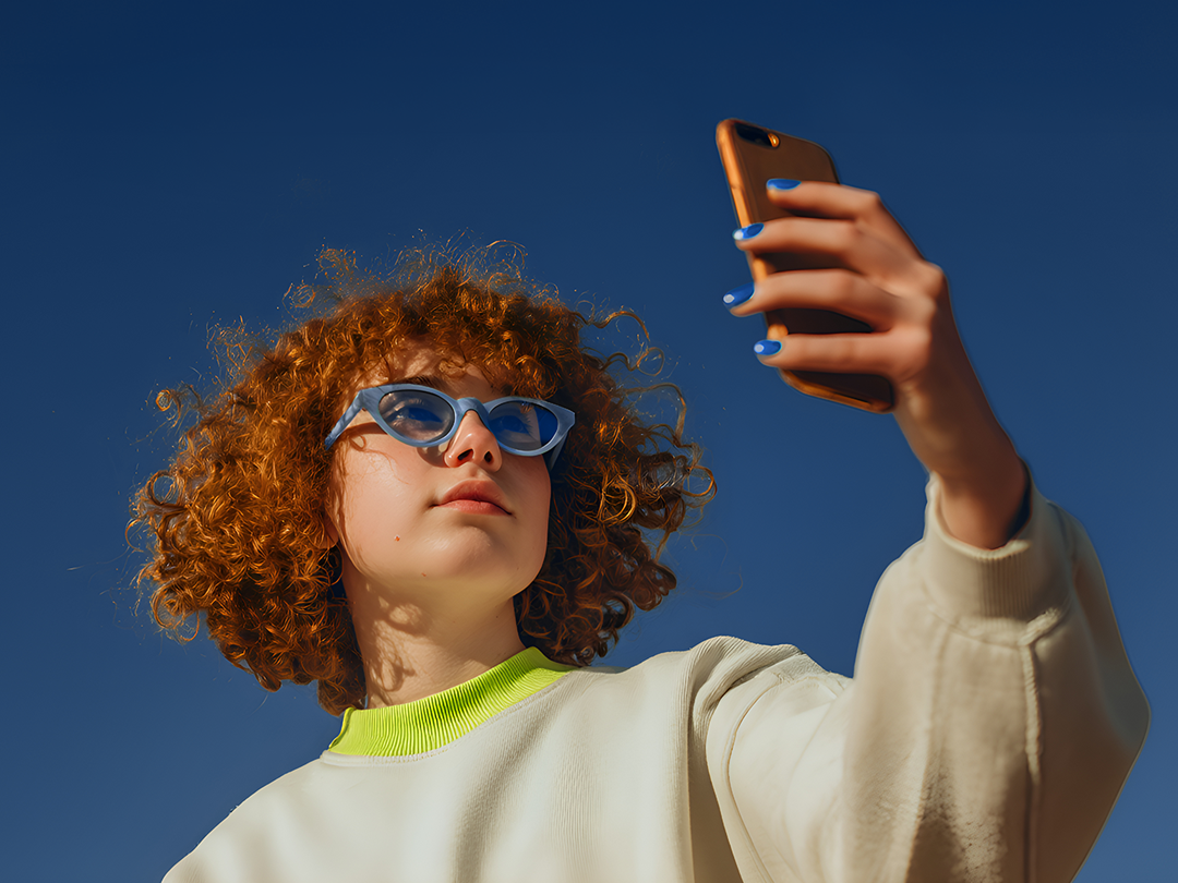 Nastolatka z rudymi, kręconymi włosami w jasnej bluzie i niebieskich okularach przeciwsłonecznych robi selfie telefonem na tle czystego, niebieskiego nieba.