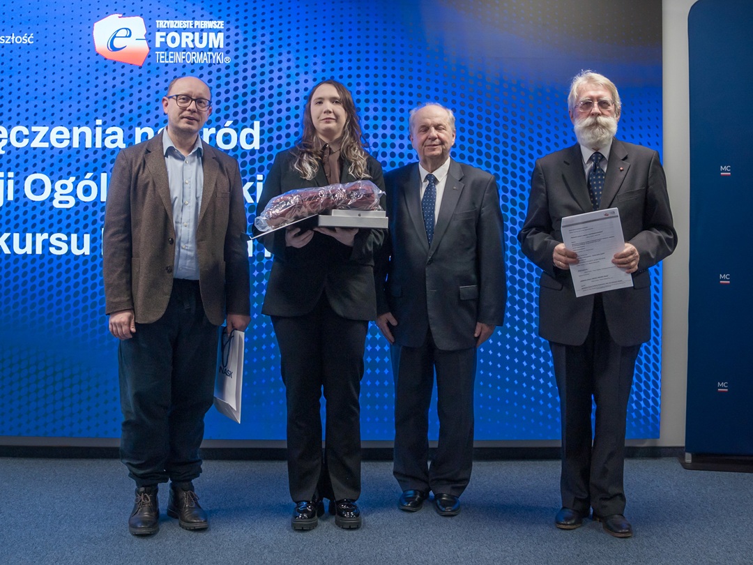 Cztery osoby stojące obok siebie na scenie podczas wydarzenia. Kobieta w środku trzyma nagrodę zapakowaną w przezroczystą folię. Trzej mężczyźni obok niej trzymają dokumenty lub torby. W tle niebieska ścianka z napisem „Forum Teleinformatyki” i wzorem kropek.